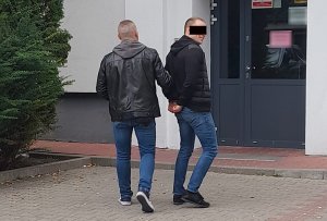 Po lewej stronie idzie nieumundurowany policjant z mokotowskiego wydziału do walki z przestępczością przeciwko mieniu. Funkcjonariusz prowadzi znajdującego się po prawej stronie podejrzanego mężczyznę, który jest odwrócony tyłem do obiektywu.