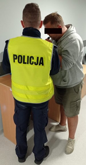 Z lewej strony umundurowany funkcjonariusz ruchu drogowego obsługujący stacjonarne urządzenie do badania trzeźwości. Z prawej strony zatrzymany mężczyzna, który dmucha w ustnik aklometru.