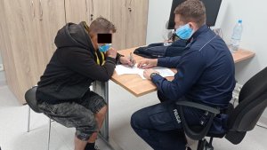 Po lewej stronie na krześle przy biurku siedzi jeden z zatrzymanych. Mężczyzna ma kajdanki założone na ręce z przodu. Podpisuje protokół zatrzymania. Po prawej stronie przed komputerem siedzi umundurowany policjant, który wykonuje czynności z zatrzymanym. Policjant daje zatrzymanemu protokół do podpisania.