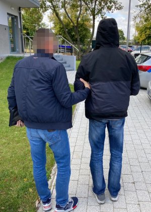 Z prawej strony zatrzymany mężczyzna. Ma on kajdanki założone z przodu. Z lewej strony nieumundurowany policjant z wydziału kryminalnego KP Warszawa Ursynów.