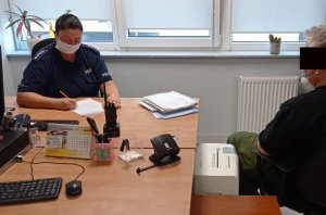 Po lewej stronie za biurkiem siedzi umundurowana policjantka. Po prawej stronie na krześle siedzi poszukiwany mężczyzna.