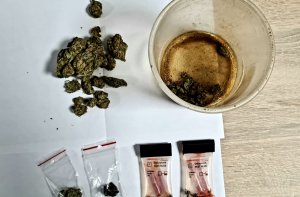 Na zdjęciu plastikowy pojemnik z zawartością brązowo-zielonej substancji, 
4 testery narkotykowe i 2 foliowe woreczki z zapięciem strunowym z zawartością zielono-brunatnego suszu oraz brązową, zbrylona masą
