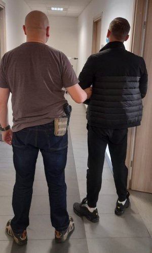 Z lewej strony zdjęcia nieumundurowany policjant mokotowskiego wydziału do walki z przestępczością przeciwko mieniu. Funkcjonariusz prowadzi znajdującego się z prawej strony 36-latka podejrzanego o kradzież z włamaniem na konto osobiste pokrzywdzonego. Mężczyzn a ma kajdanki założone na ręce z przodu.