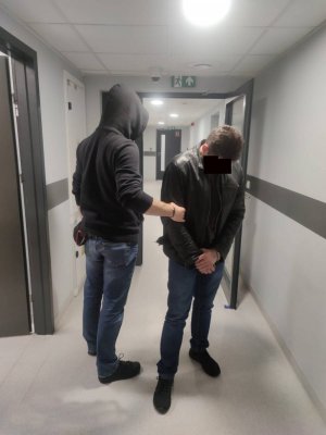Po lewej stronie nieumundurowany policjant w WWP KP Warszawa Ursynów doprowadza do pomieszczeń dla osób zatrzymanych podejrzanego mężczyznę. Z prawej strony  28-latek podejrzany o posiadanie narkotyków. Mężczyzna ma kajdanki założone na ręce z przodu.
