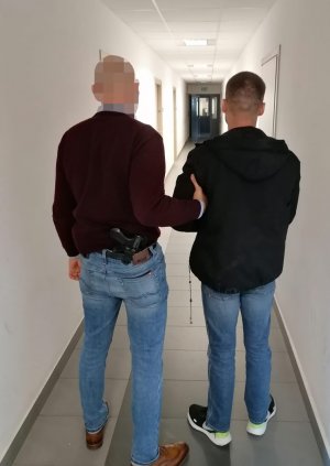 Z lewej strony zdjęcia nieumundurowany policjant mokotowskiego wydziału do walki z przestępczością przeciwko życiu i zdrowi.. Funkcjonariusz prowadzi poszukiwanego 34-latka znajdującego się z prawej strony Mężczyzn a ma kajdanki założone na ręce z przodu.