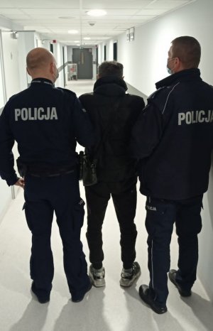 Z lewej i z prawej strony na zdjęciu umundurowani policjanci z Komisariatu Policji Warszawa Ursynów. Funkcjonariusze prowadzą zatrzymanego 50-latka podejrzanego o kradzieże paliwlia Mężczyzna ma kajdanki założone na ręce zapięte z przodu.