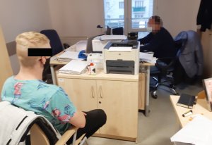 Z lewej strony przed biurkiem na krześle siedzi mężczyzna podejrzany o kradzieże z włamaniem Na przeciwko niego, po prawej stronie na zdjęciu, za biurkiem siedzi nieumundurowany policjant, która wykonuje czynności z podejrzanym.