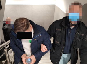 Z prawej strony nieumundurowany policjant z WK. Z lewej strony zatrzymany mężczyzna. Poszukiwany ma kajdanki założone na ręce z przodu .
