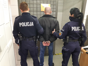 Z lewej strony umundurowany policjant, z prawe strony umundurowana policjanta z wydziału wywiadowczo-patrolowego. W środku zatrzymany mężczyzna podejrzany o oszustwo. Mężczyzna ma kajdanki założone na ręce z tyłu.