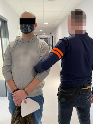 Z lewej strony 33-letni mężczyzna podejrzany o zmuszanie do określonego zachowania groźby karalne. Mężczyzna ma kajdanki założone na ręce z przodu. Z prawej strony nieumundurowany policjant z ursynowskiego wydziału kryminalnego.