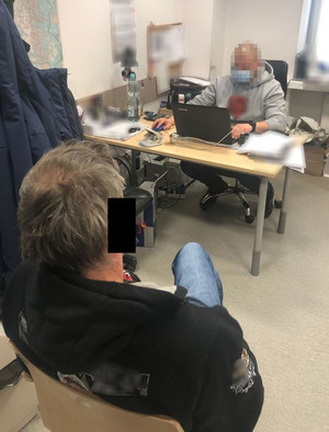 Z lewej strony zdjęcia podejrzany mężczyzna siedzi na krześle. Ma kajdanki założone na ręce z przodu. Na przeciwko niego za biurkiem siedzi nieumundurowany policjanci wykonuj ący czynności z zatrzymanym.