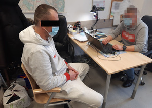 Z lewej strony zatrzymany mężczyzna siedzi na krześle obok biurka. Ma kajdanki założone na ręce z przodu. Z prawej strony nieumundurowany policjant, wykonuje czynności z z jego udziałem.