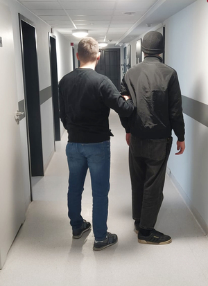 Z lewej strony nieumundurowany policjant. Z prawej strony zatrzymany mężczyzna. Zdjęcie zrobione na korytarzu w policyjnym areszcie.