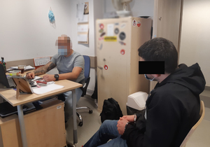 Z lewej strony za biurkiem nieumundurowany policjant wykonuje czynności z siedzącym po prawej stronie na krześle zatrzymanym 35-latkiem. Mężczyzna ma kajdanki założone na ręce z przodu.