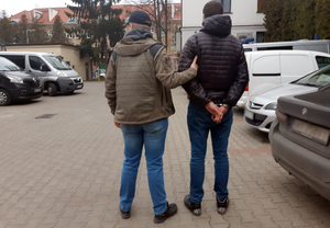 Z prawej strony nieumundurowany policjant prowadzi znajdującego się z lewej strony zatrzymanego mężczyznę. Zatrzymany ma kajdanki założone na ręce ułożone do tyłu.