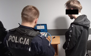 Z prawej strony przy urządzeniu do badania trzeźwości stoi umundurowany policjant, który przygotowuje urządzenie do przeprowadzenia próby trzeźwości wobec stojącego z prawej strony zatrzymanego 22 latka.