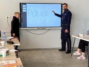 W klasie przy tablicy multimedialnej przed uczniami stoi umundurowany policjant, który prowadzi warsztaty edukacyjne z dziećmi. Na przeciwko niego w ławkach siedzą słuchacze.