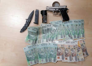 Na zdjęciu pistolet koloru srebrnego z czarną rękojeścią, magazynek do pistoletu, czarny rozkładany scyzoryk, gaz pieprzowy, banknoty 100 złotowe i 200 złotowe.