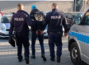 Z lewej strony oraz prawej  strony na zdjęciu umundurowani policjanci. Funkcjonariusze prowadzą znajdującego się między nimi zatrzymanego mężczyznę. Podejrzany ma kajdanki założone na ręce do tyłu.