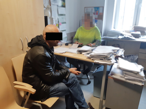 Z lewej strony na krześle siedzi mężczyzna podejrzy o kradzież.  Z prawej strony za biurkiem siedzi nieumundurowana policjantka, która wykonuje czynności z jego udziałem.