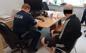 Z lewej strony umundurowany policjant siedzi przy biurku i sporządza dokumentację w związku z zatrzymaniem poszukiwanego. S prawej strony siedzi poszukiwany mężczyzna. Ma on kajdanki założone na ręce ułożone do przodu.