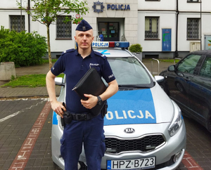 Umundurowany policjant na tle oznakowanego radiowozu policyjnego oraz budynku Komendy Rejonowej Policji Warszawa II