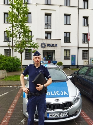 Umundurowany policjant na tle oznakowanego radiowozu policyjnego oraz budynku Komendy Rejonowej Policji Warszawa II