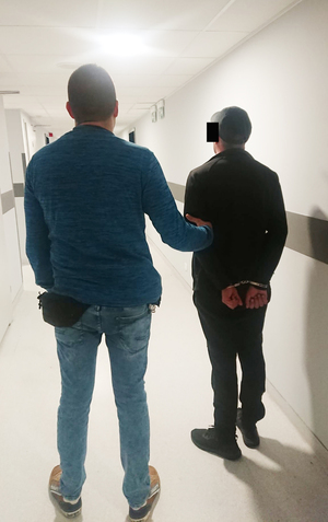 Z prawej strony nieumundurowany policjant. Z lewej strony mężczyzna podejrzany o posiadanie narkotyków. Policjant prowadzi go do policyjnej celi. Zatrzymany mężczyzna ma kajdanki założone na ręce złożone do tyłu.