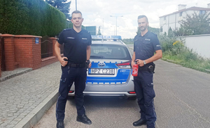 Z lewej i prawej strony stoją umundurowani policjanci. Funkcjonariusz z prawej strony ma gaśnicę w ręku. Między nimi stoi oznakowany radiowóz.