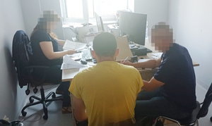 Z lewej strony za biurkiem siedzi nieumundurowana policjantka. Pośrodku w żółtej podkoszulce siedzi podejrzany zwrócony przodem do policjantki. Z prawej strony obok podejrzanego siedzi tłumacz języka gruzińskiego.