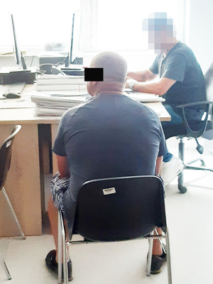 Na pierwszym planie siedzi zatrzymany mężczyzna, z którego udziałem policjant wykonuje czynności. Policjant siedzi za biurkiem i przesłuchuje podejrzanego.