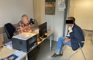 Z lewej strony za biurkiem siedzi nieumundurowany policjant wykonujący czynności z zatrzymanym mężczyzną. Z prawej strony przed biurkiem siedzi podejrzany mężczyzna