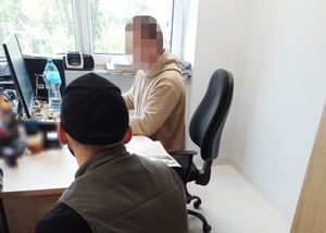 Z lewej strony przy biurku siedzi mężczyzna podejrzany o kradzież. Z prawej strony za biurkiem siedzi nieumundurowany policjant, który wykonuje czynności z zatrzymanym mężczyzną.