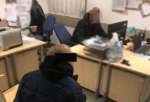 Z lewej strony na krześle siedzi podejrzany mężczyzna, ma kajdanki założone na ręce z tyłu. Z prawej strony za biurkiem siedzi nieumundurowany policjant, który wykonuje czynności z podejrzanym. Policjant sporządza dokumentację.