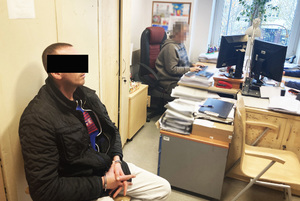 Z lewej strony na zdjęciu na krześle siedzi podejrzany mężczyzna. Ma kajdanki założone na ręce ułożone do przodu. Dalej w głąb zdjęcia lekko po prawe stronie siedzi nieumundurowana policjanta, która przesłuchuje podejrzanego.