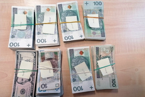 Na zdjęciu widać 8 plików gotówki ułożonej w nominały. 6 plików składa się z banknotów 100 złotowych, jednym są banknoty 10, 20 i 50 złotowe i w jednym dolary amerykańskie z pięciodolarowym banknotem na wierzchu.