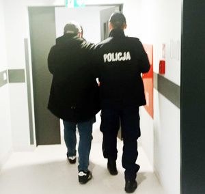 Z lewej strony na zdjęciu zatrzymany mężczyzna. Ma on kajdanki założone ręce ułożone do przodu. Obok niego z prawej strony zdjęcia umundurowany policjant, który prowadzi zatrzymanego korytarzem w komisariacie Policji.