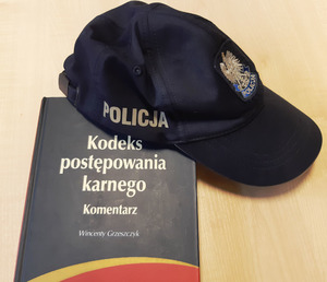 Na podłożu leży książka z napisem Kodeks postępowania kartnego -  komentarz oraz czapka z napisem Policja.