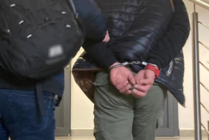 Policjant prowadzi po schodach zatrzymanego mężczyznę, który ma założone kajdanki na ręce z tyłu.