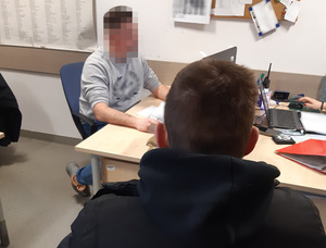 Z lewej strony za biurkiem siedzi nieumundurowany policjant, który przesłuchuje siedzącego z prawej strony mężczyznę, podejrzanego o kradzież sklepową.