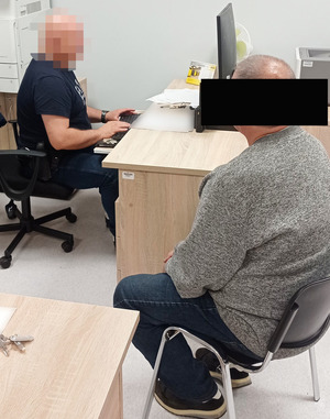 Nieumundurowany policjant siedzi przy biurku i przesłuchuje zatrzymanego mężczyznę. Zatrzymany siedzi przy biurku, ma założone kajdanki na ręce.