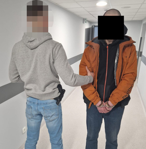 Nieumundurowany policjant prowadzi zatrzymanego mężczyznę. Na ręce trzymane z przodu ma założone kajdanki.