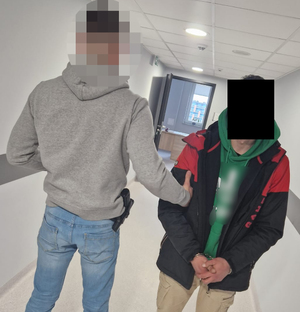Nieumundurowany policjant prowadzi zatrzymanego mężczyznę. Na ręce trzymane z przodu ma założone kajdanki.