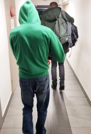 Mężczyzna ubrany w zielona bluzę z kapturem stoi na korytarzu. przed nim idzie drugi mężczyzna,.