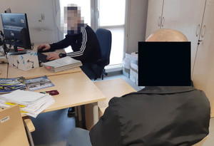 Policjant siedzi przy biurku i wykonuje czynności z zatrzymanym. Drugi mężczyzna siedzi obok biurka. Na dłoniach ma założone kajdanki.