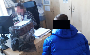 Policjant nieumundurowany siedzi przy biurku i wykonuje czynności z zatrzymanym mężczyzną, który siedzi obok biurka. Ubrany w niebieską kurtkę.