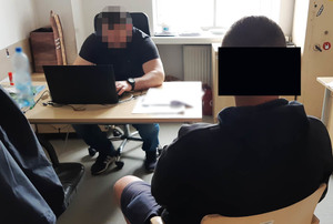 Policjant nieumundurowany wykonuje czynności przy biurku z zatrzymanym mężczyzną.