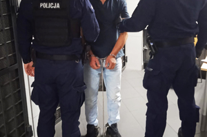 Dwóch umundurowanych policjantów prowadzi zatrzymanego mężczyznę, który ma założone kajdanki na ręce trzymane z przodu.
