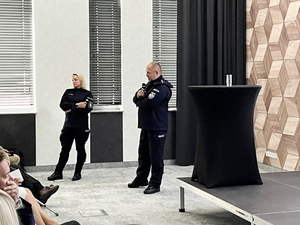 Umundurowana policjantka z policjantem stoją z mikrofonami w rękach i prowadzą wykład.
