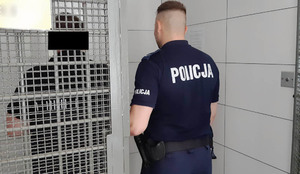 Umundurowany policjant trzyma za łokieć mężczyznę. Obaj stoją w pomieszczeniu z kratami.